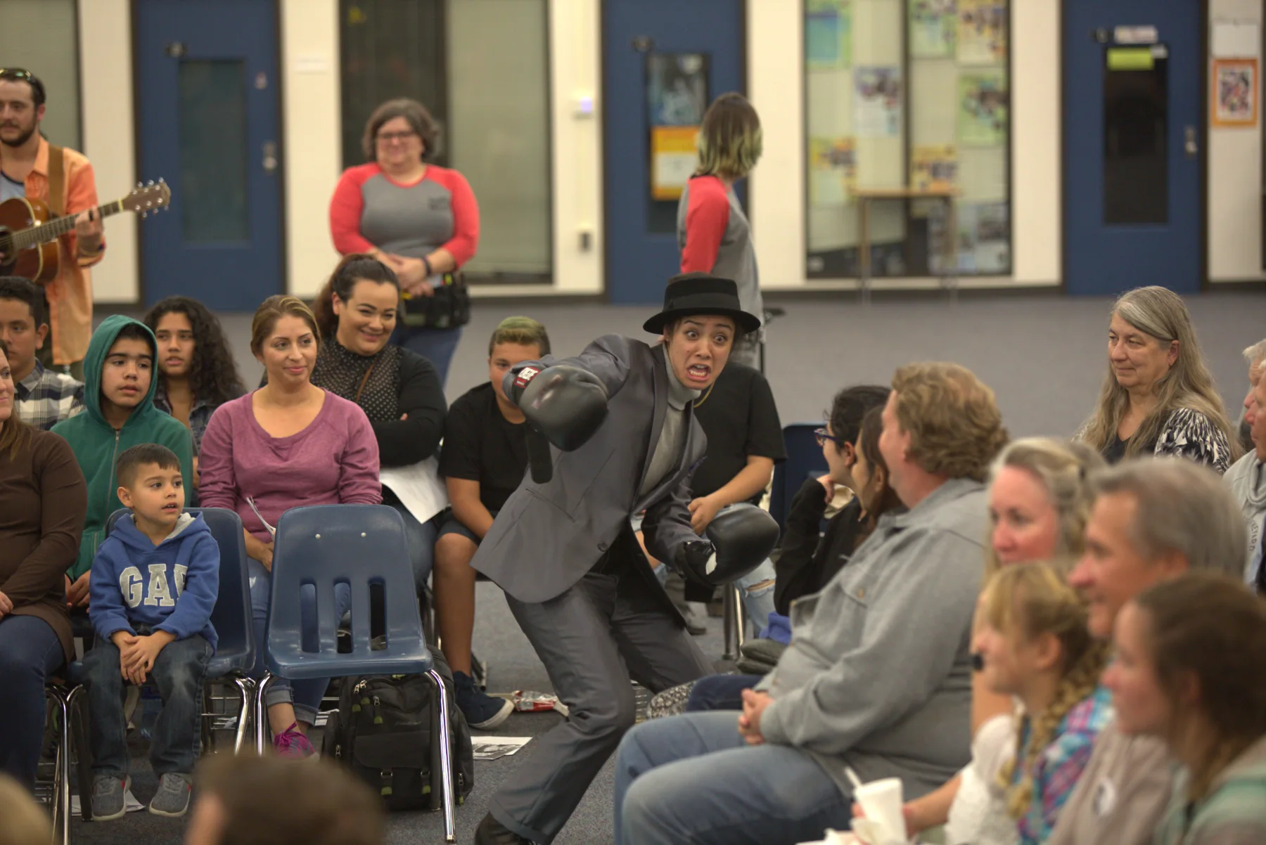 A performer in a hat and boxing gloves mid-scene, the audience seated close around her in a community room