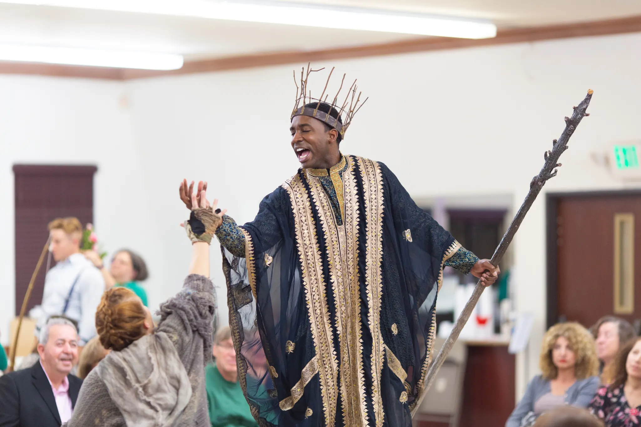 A performer in a twig crown mid-speech, hands reaching up from the audience around him